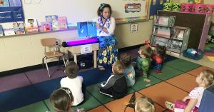 Preschool teacher leads a storytime as children sit on a colorful classroom rug listening to a blue book read aloud.