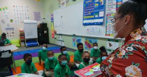 TheFutureofSchools_featuredimage - Mississippi First Teacher wearing a mask reads a children's book to a classroom of attentive students on a colorful rug.