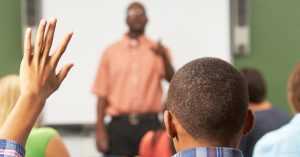 Audience in a classroom with a person raising a hand while a speaker stands at the front.