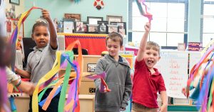 TransformingPreK_featuredimage - Mississippi First Three children in a colorful classroom waving rainbow ribbons during a dance activity.