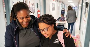 Two young women in a school hallway share a comforting, supportive moment with smiles.