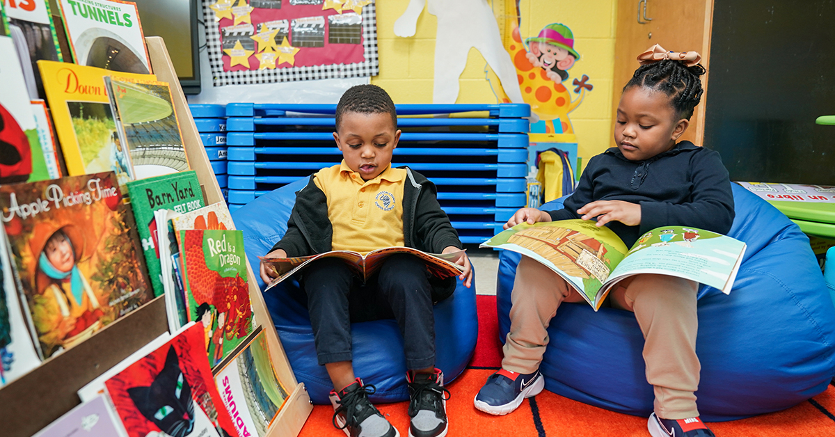 Two young children sit on blue bean bags reading colorful picture books in a bright children’s reading corner with books nearby.